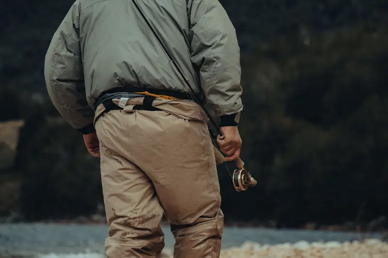 An adult man in fishing gear holding a fly rod near a scenic river outdoors
