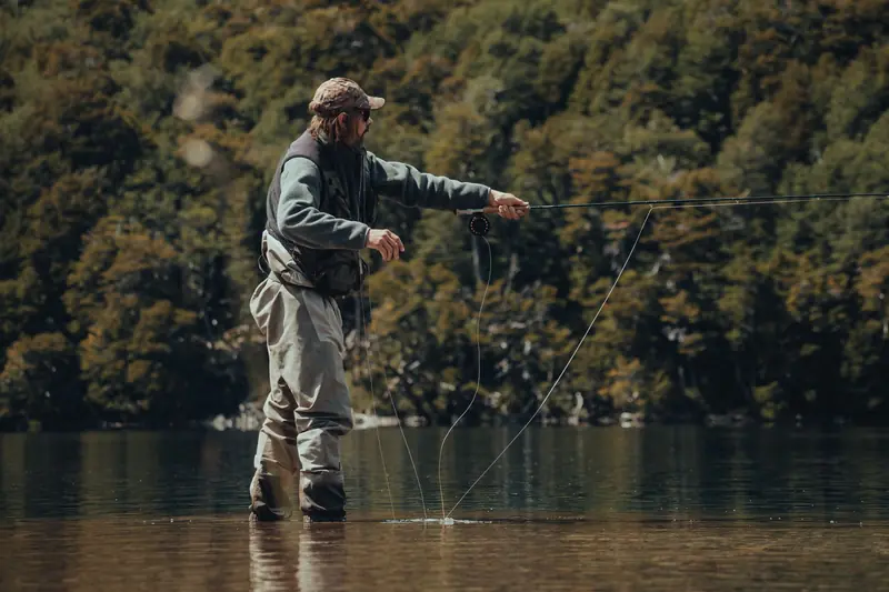 A man in waders fly fishing in a tranquil lake surrounded by lush green forest